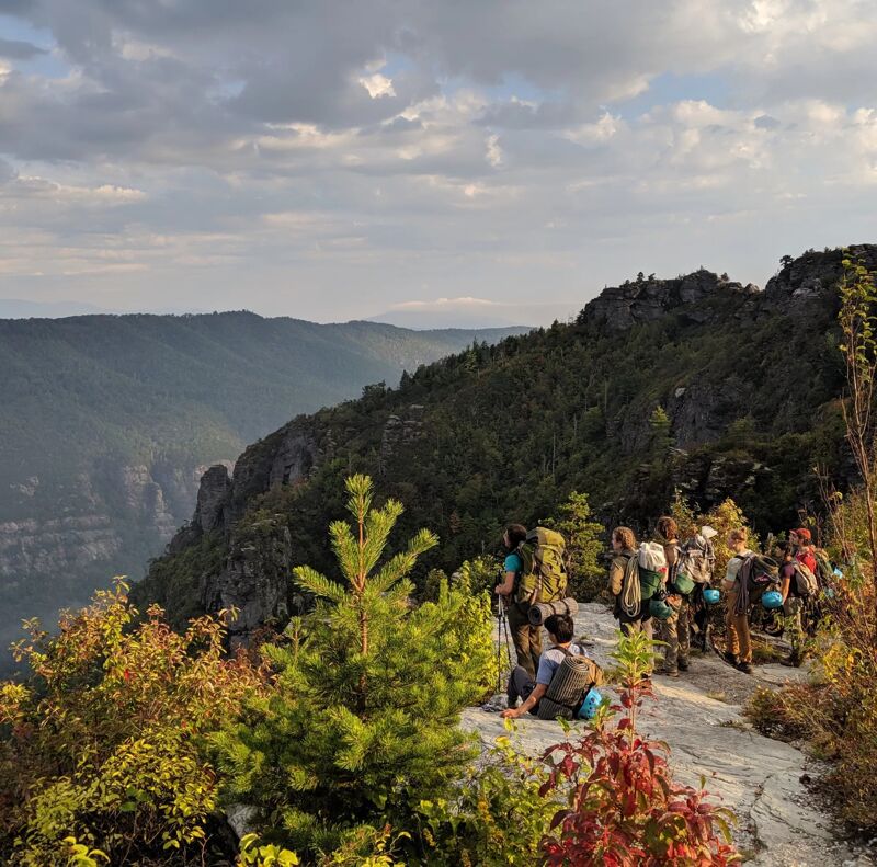 A group of hikers is walking along a rocky trail on a mountain ridge. They are carrying backpacks and hiking poles. The trail is surrounded by trees and shrubs. In the distance, there are mountains and a valley. The sky is cloudy and the sun is shining. It looks like a beautiful day for a hike.